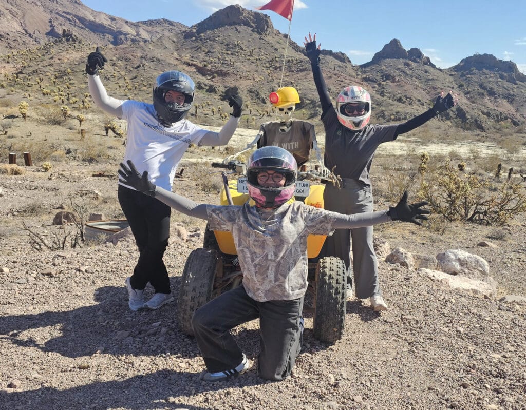 Four friends in helmets pose by a yellow ATV amid Nevada desert hills on a Las Vegas RZR off-road tour near the Colorado River.