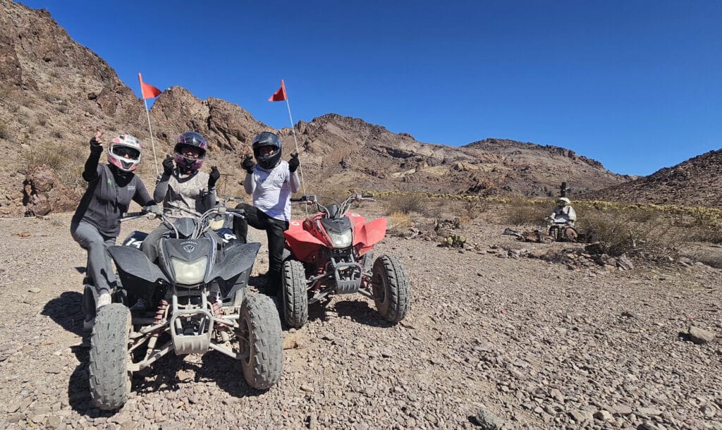 Adventurers in helmets ride ATVs on Nevadas rocky trails near Las Vegas, with red flags waving—perfect for ATV tours or RZR off-road fun!.