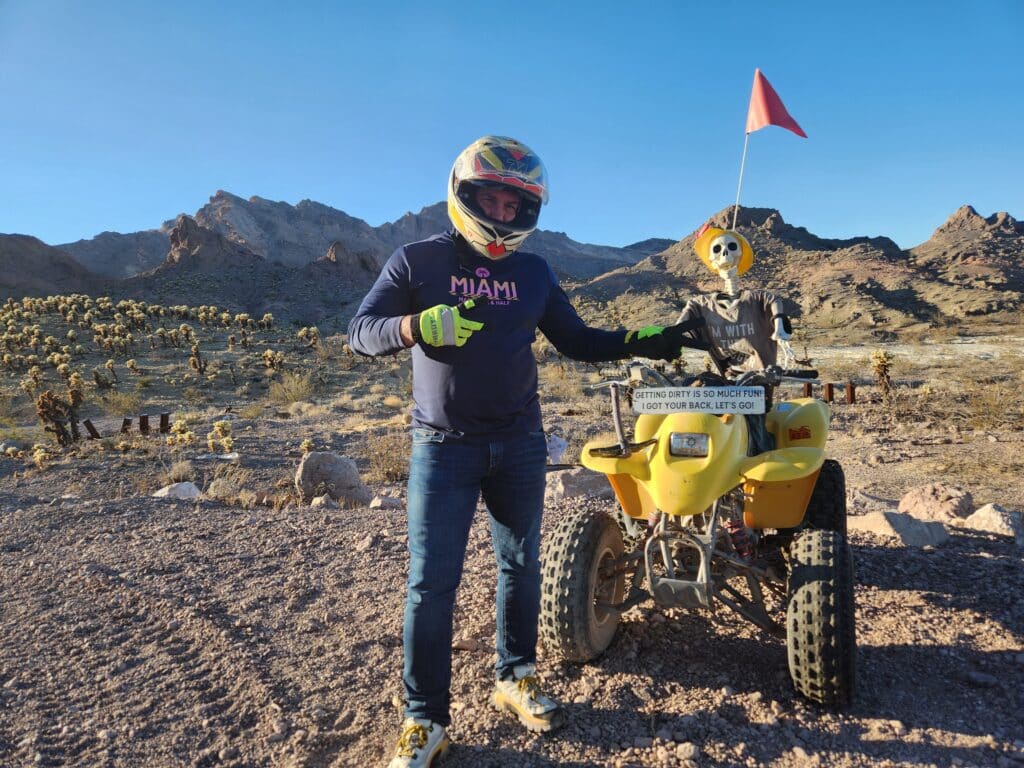 Adventurer in helmet and Miami shirt stands by yellow ATV with skeleton near Nevada desert, cacti, and Colorado River views.