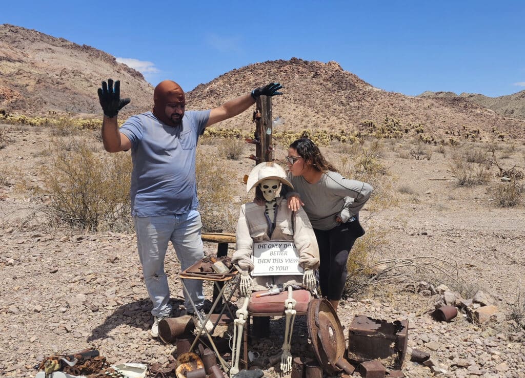 Man with raised hands and woman pose by skeleton with sign in Nevada desert near Las Vegas; perfect for Ghost Town sightseeing.
