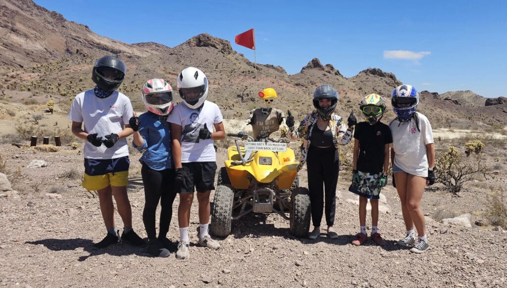 Six adventurers in helmets pose by a yellow ATV in Nevada’s desert near Las Vegas on an exciting RZR off-road tour.