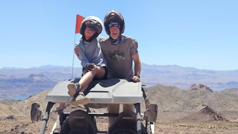 Smiling adventurers in helmets enjoy a RZR off-road ride near Las Vegas, Nevada, with desert views and a red flag flying high.