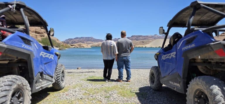 A couple holds hands by a scenic Nevada lake, flanked by blue Polaris RZRs—perfect for Las Vegas ATV tours near the Colorado River.