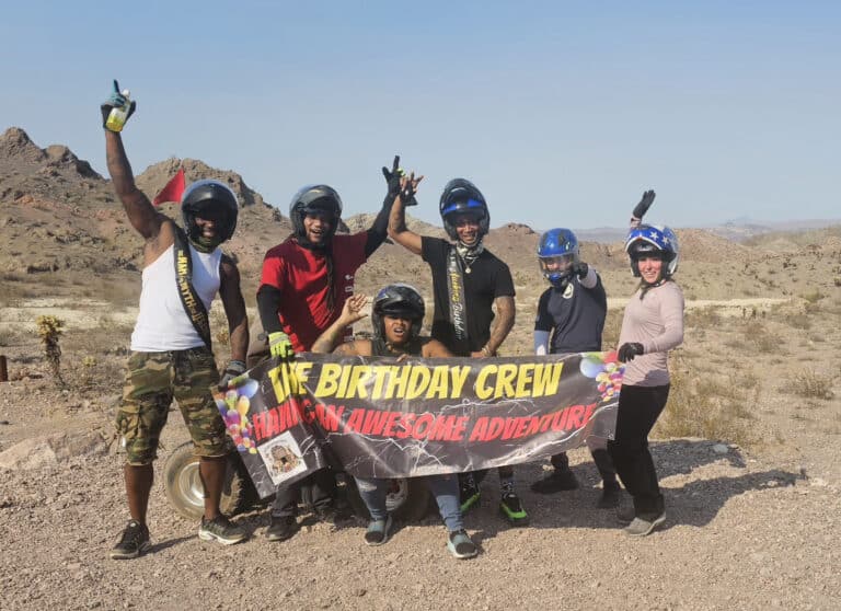 Six friends in helmets pose in Nevada’s rocky desert after a Las Vegas RZR tour, holding a “Birthday Crew Adventure” banner.