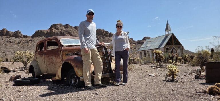 Couple poses by a rusty vintage car in a Nevada desert ghost town near Las Vegas, with rocky hills and old church—perfect for ATV tours.