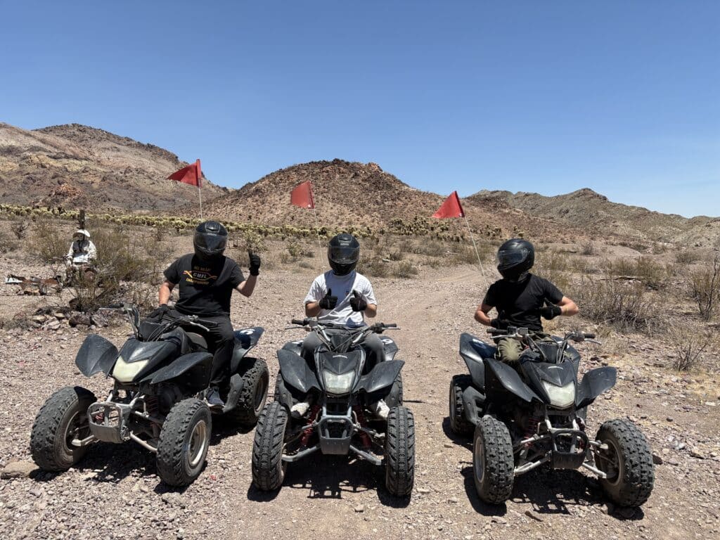 Explore Nevada’s rocky desert near Las Vegas on ATV tours—three riders with red flags, mountains, and dry brush in the background.