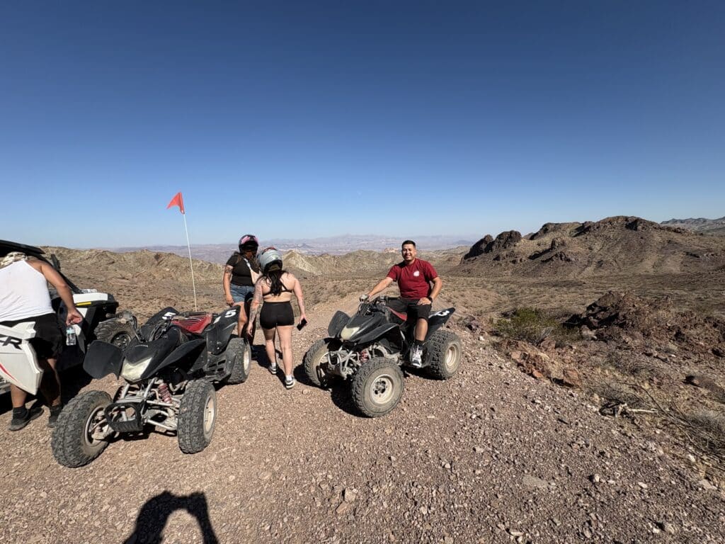 Adventurers with helmets pose by ATVs on a rocky hilltop near Las Vegas, Nevada, overlooking desert and mountain views.