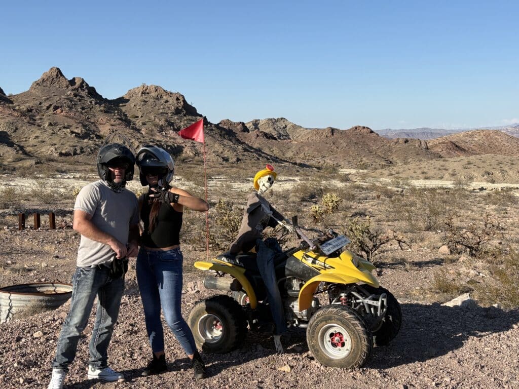 Two riders in helmets pose by a yellow ATV with a helmeted skeleton near Las Vegas, Nevada—perfect for ATV tours or Ghost Town rides.