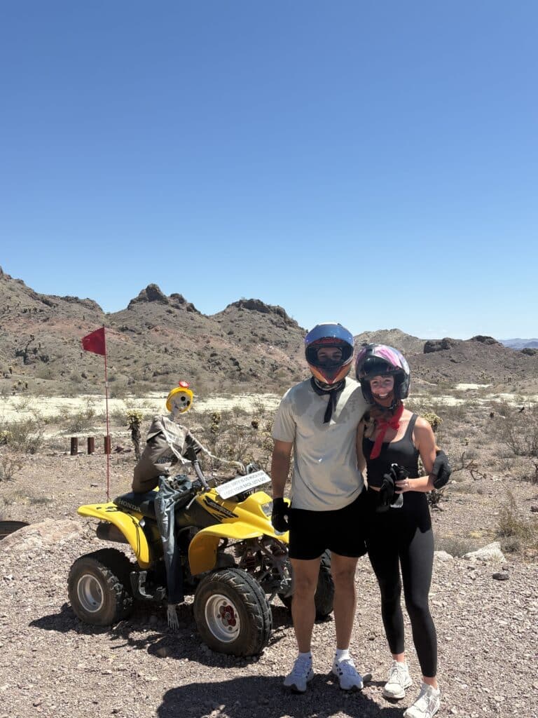 Smiling couple in helmets pose by a yellow ATV and skeleton near Las Vegas on a Nevada RZR off-road desert tour.