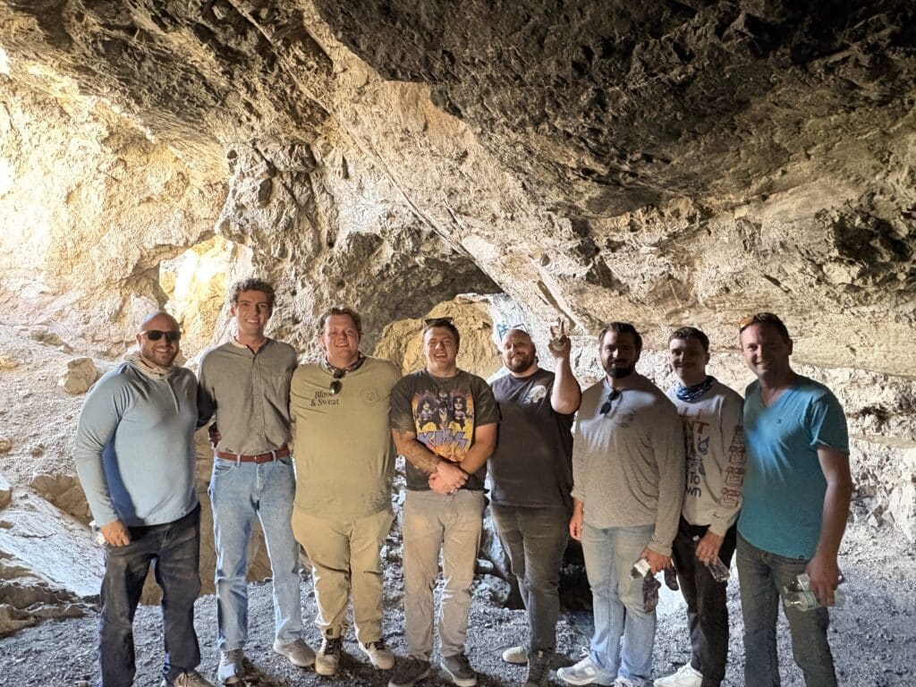 Eight men smile inside a rocky Nevada cave near Las Vegas, lit by sunlight during a Colorado River ATV and RZR off-road adventure.