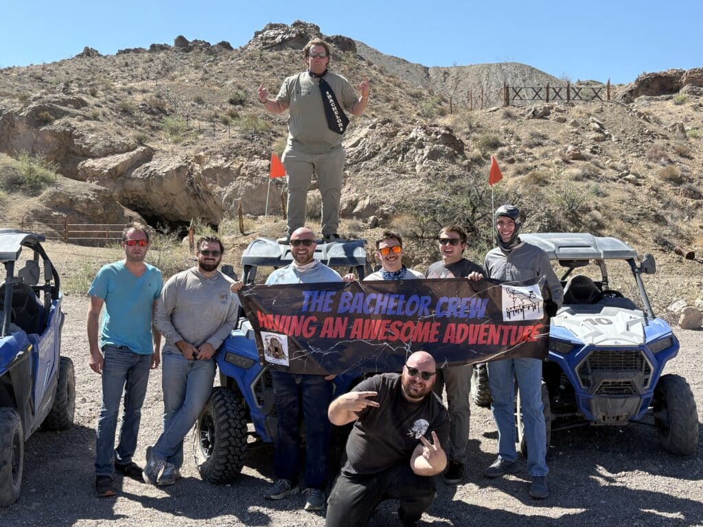 Eight men pose by ATVs in Nevada near the Colorado River, holding a Bachelor Crew banner after an epic RZR off-road adventure.