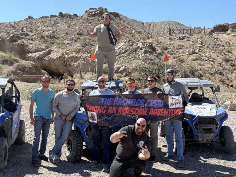 Eight men pose by ATVs in Nevada near the Colorado River, holding a Bachelor Crew banner after an epic RZR off-road adventure.
