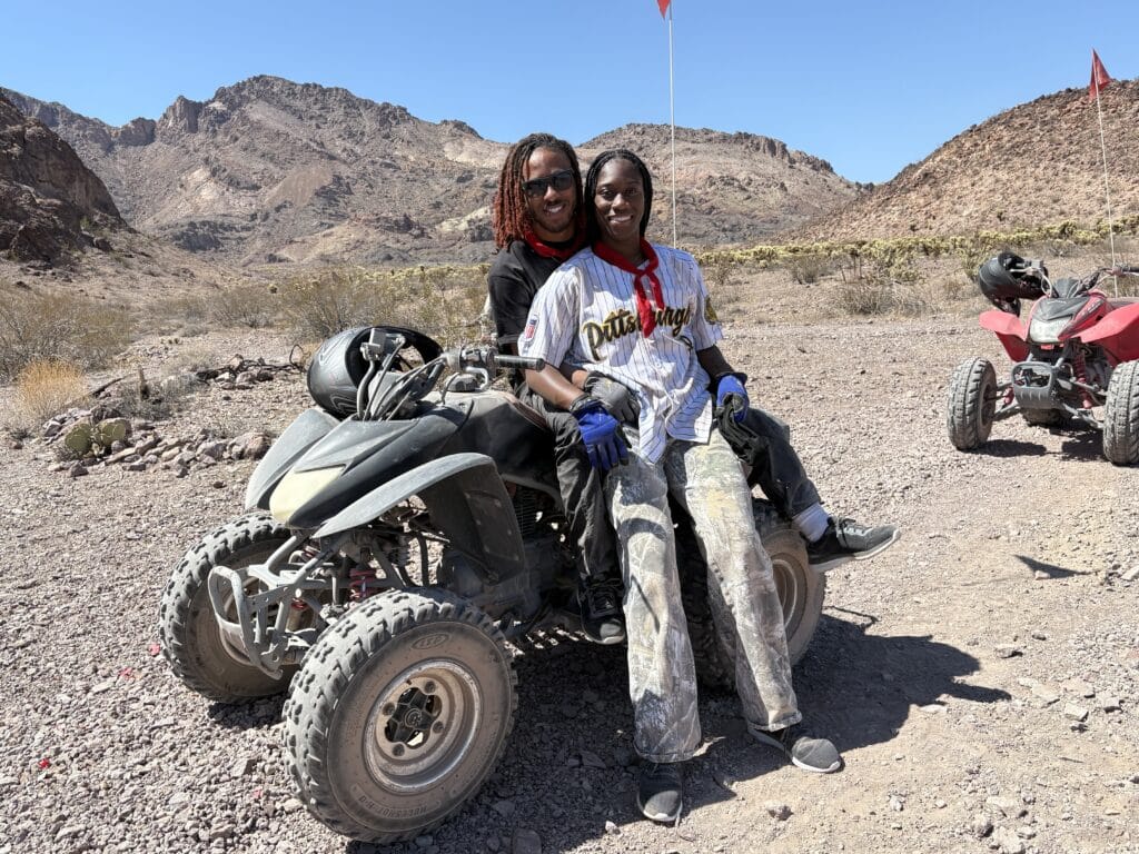 Smiling duo on Las Vegas ATV tour in Nevada desert near the Colorado River, with mountains and Ghost Town sightseeing views.