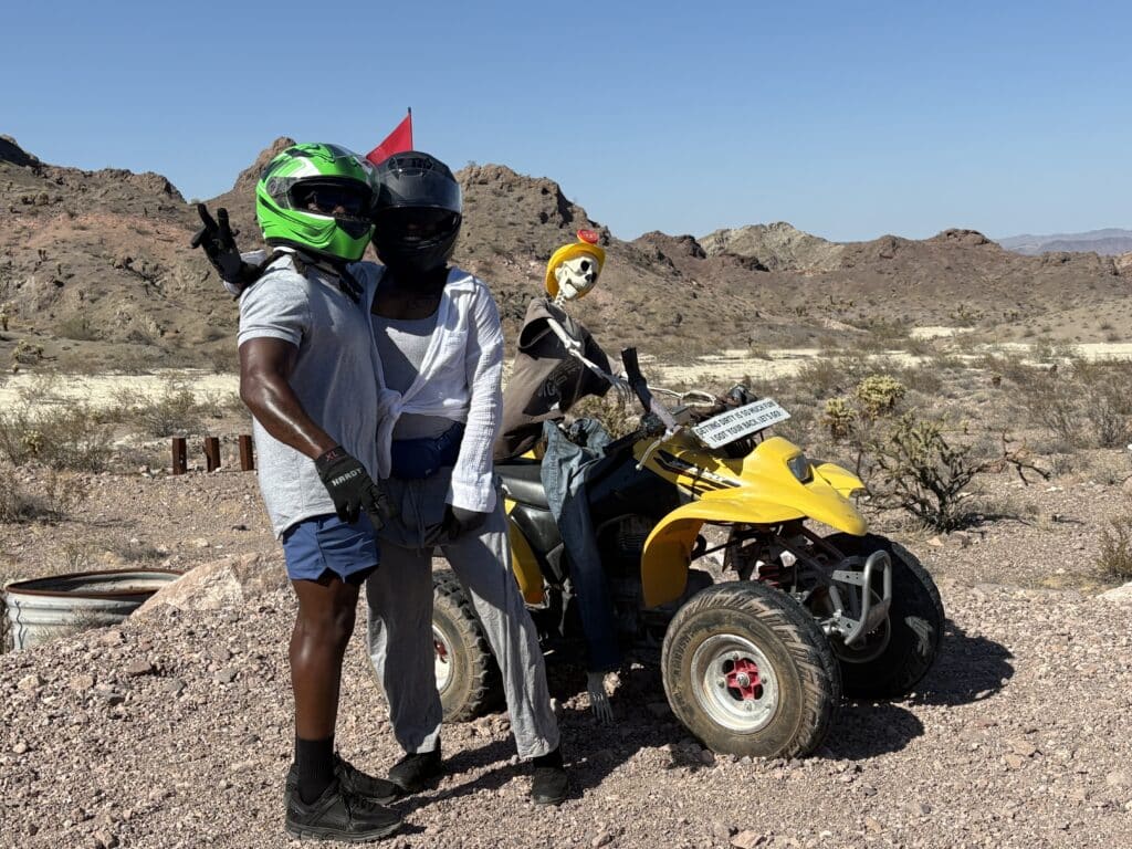 Two riders in helmets pose by a yellow ATV and skeleton near Las Vegas, Nevada—perfect for ATV tours & Ghost Town sightseeing.