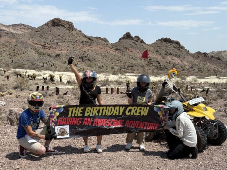 Four riders in helmets pose with a Birthday Crew banner during a Las Vegas ATV tour near the Colorado River in Nevada desert.