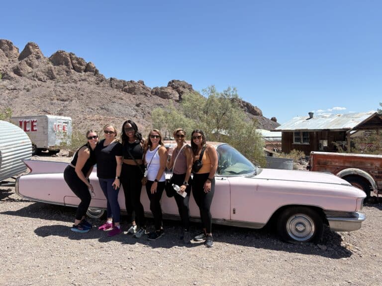 Six women smile by a vintage pink car in a Nevada ghost town near Las Vegas, perfect for RZR tours and Colorado River adventures.