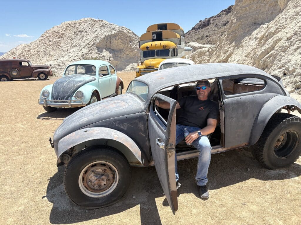 A man in sunglasses and a cap sits in a rusty VW Beetle near vintage cars and school buses in Nevada’s desert—perfect for Ghost Town tours.