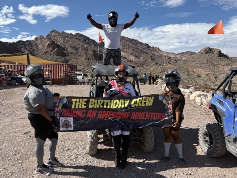Adventurers in off-road gear hold a Birthday Crew banner by an ATV in Nevada, with Rocky mountains and blue sky behind.
