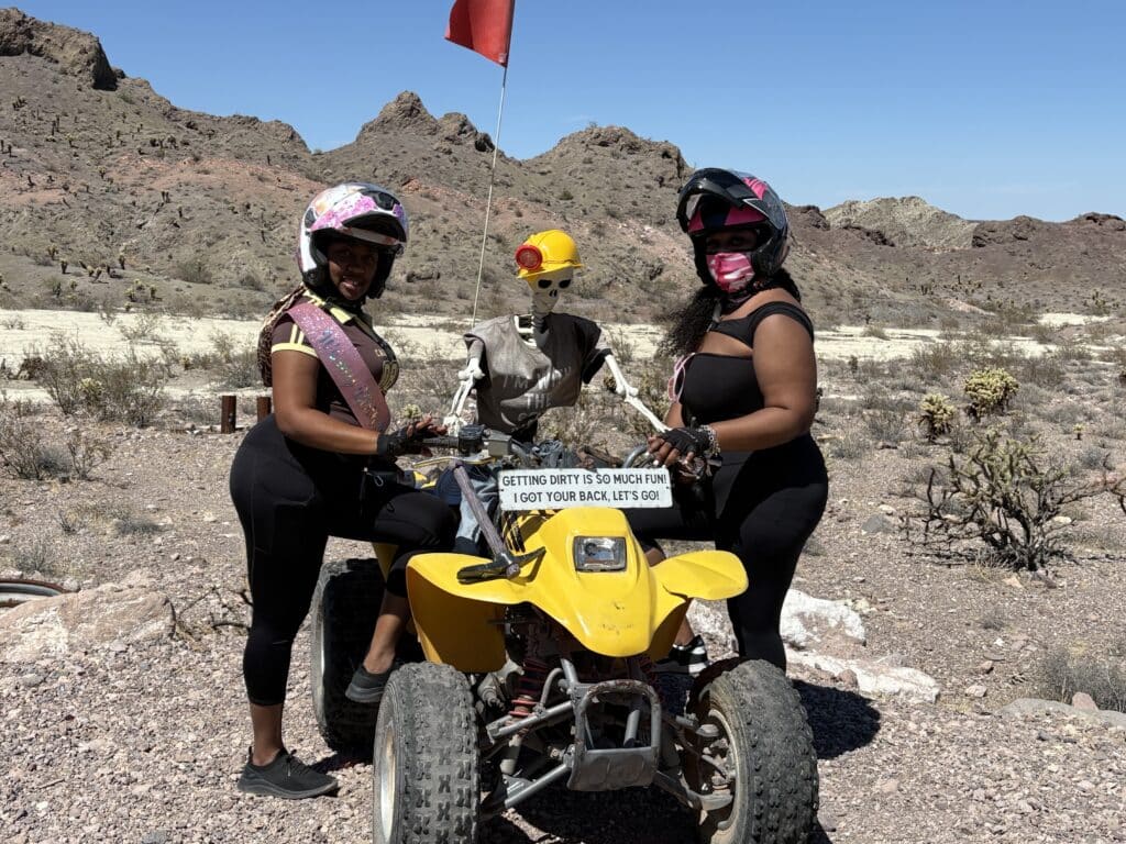 Two women in helmets pose by a safety-clad skeleton on an ATV near Las Vegas, Nevada—perfect for ATV tours & RZR off-road fun!.