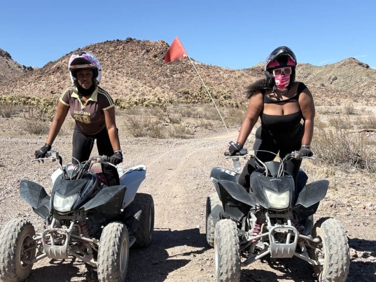 Two women enjoy an ATV tour near Las Vegas, Nevada, riding black ATVs on rocky desert trails with mountains and dry shrubs nearby.