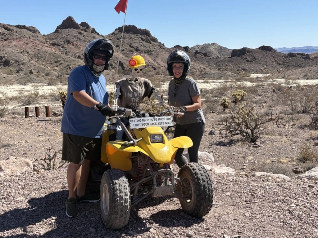 Two riders in helmets pose by a yellow ATV & skeleton near Nevada’s desert, perfect for Las Vegas RZR tours & Ghost Town rides.