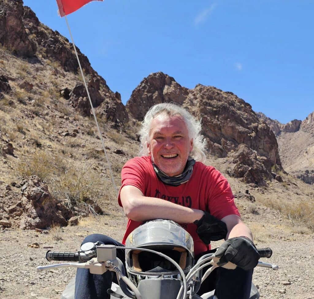 Smiling man in red shirt & gloves sits on motorcycle near Las Vegas, Nevada, with desert mountains and Colorado River views.