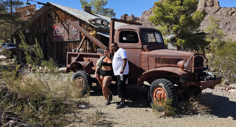 Smiling couple by vintage tow truck at Nevada ghost town near Las Vegas, with Coca-Cola sign and desert hills—ideal sightseeing spot.