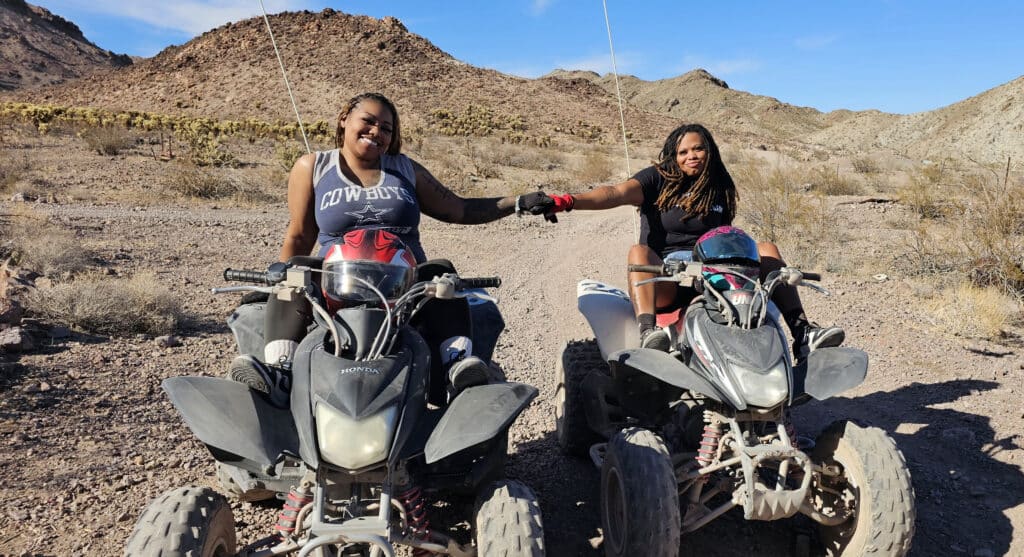 Two women hold hands on ATVs in the Nevada desert near Las Vegas, enjoying ATV tours and off-road adventures by the Colorado River.