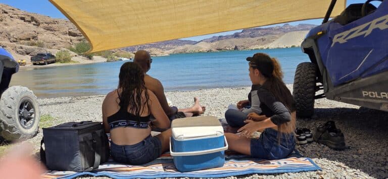 Three people relax lakeside near Las Vegas on the Colorado River—shaded picnic, ATVs, RZR rides, and Nevada mountain views.