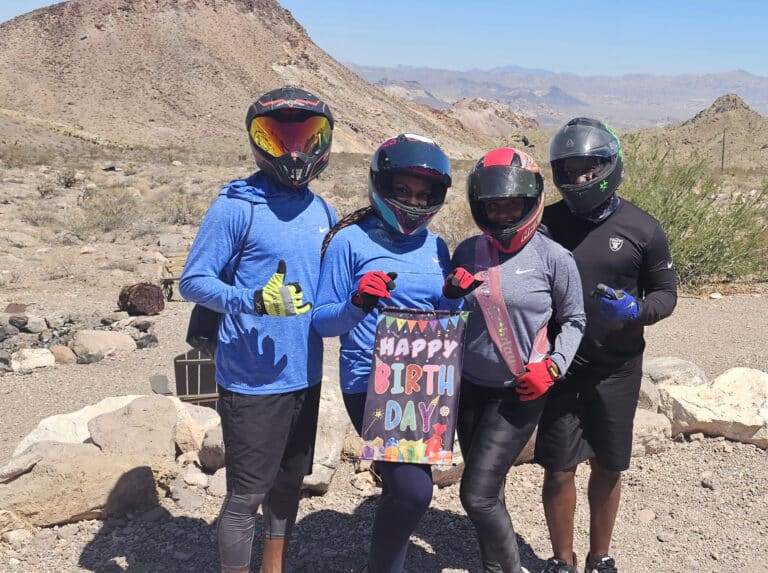 Four friends in helmets and sunglasses celebrate with a Happy Birthday sign during a Las Vegas ATV tour near the Colorado River.