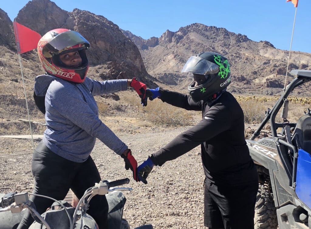 Two riders in helmets touch fingertips by ATVs in Nevada’s rocky desert—Michelangelo style—during a Las Vegas RZR tour.