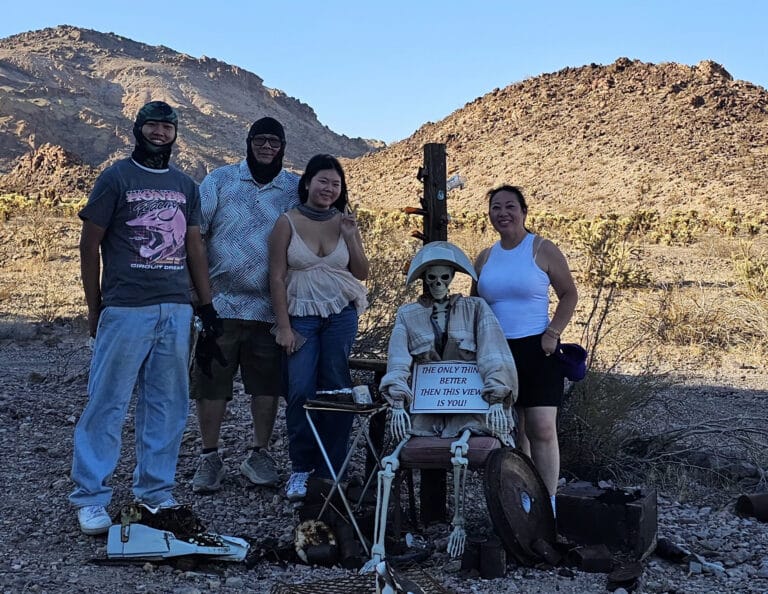Four people in Nevada’s rocky desert pose by a skeleton with a sign: The only thing deader than this view is you!—perfect for Las Vegas ATV tours.