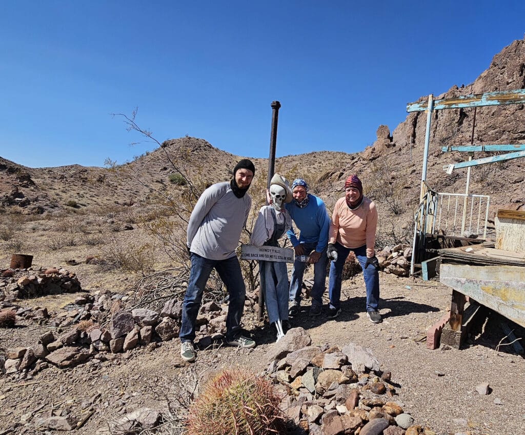 Group poses by a dressed skeleton with a sign in Nevadas desert near Las Vegas—perfect for Ghost Town sightseeing or ATV tours.