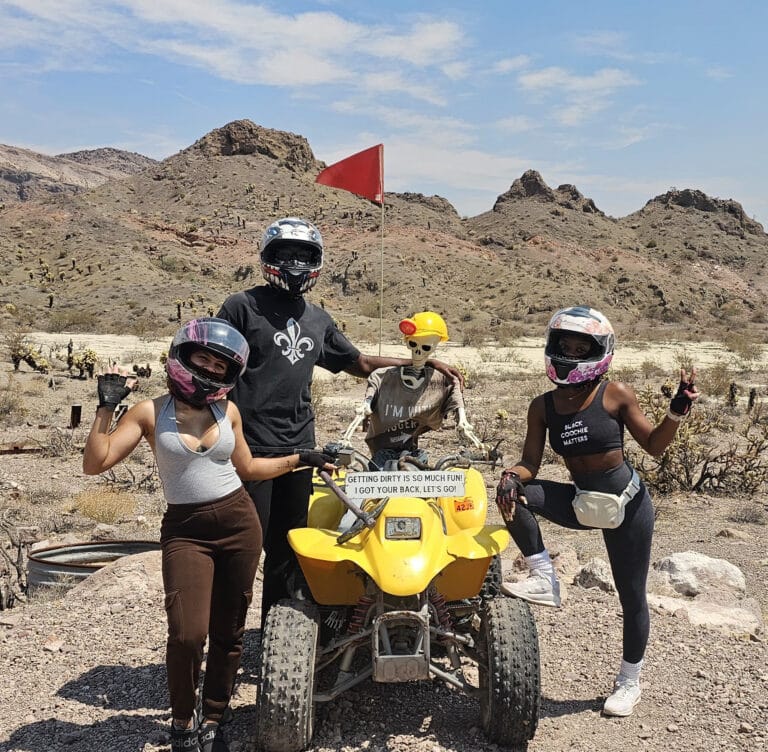 Adventurers in helmets and masks flash peace signs with a skeleton on a yellow ATV in Nevada’s rocky desert near Las Vegas.
