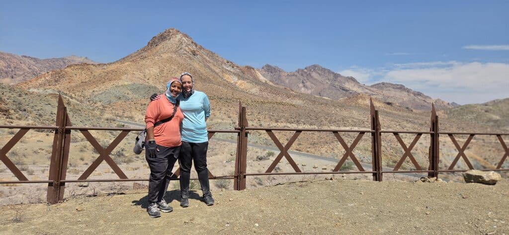 Smiling couple in outdoor gear embrace by a wooden fence near the Colorado River with Nevada mountains—perfect for ATV tours!.
