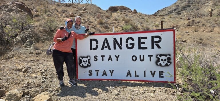 Two women in headscarves and sportswear by a “DANGER” sign in Nevada desert near Las Vegas, ideal for ATV or RZR off-road tours.