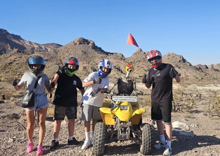 Group poses with thumbs up by yellow ATV and skeleton in Nevada desert near Las Vegas—ATV tour adventure under blue sky.