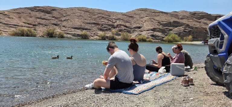 Friends picnic by the Colorado River near Las Vegas, with ducks, rocky Nevada hills, ATV tours gear, and clear blue skies.
