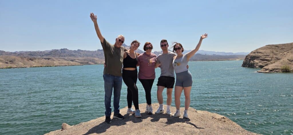 Group smiles on rocky Nevada shore by the Colorado River near Las Vegas, arms raised, mountains and clear sky behind them.