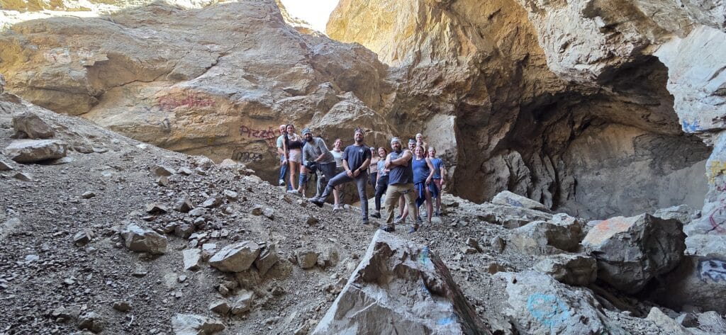 Explorers pose on rocky terrain inside a graffiti-covered cave near the Colorado River, Nevada, sunlight streaming above.