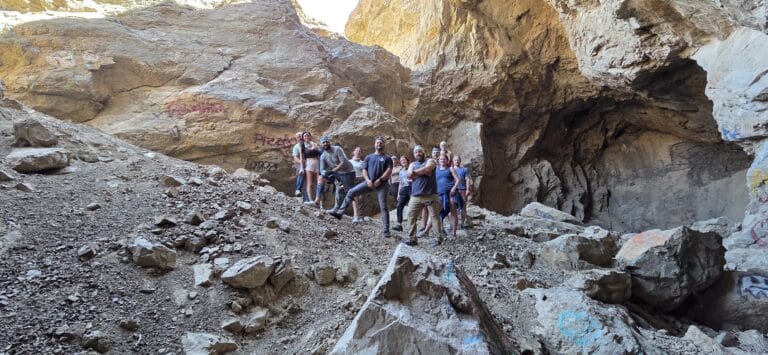 Explorers pose on rocky terrain inside a graffiti-covered cave near the Colorado River, Nevada, sunlight streaming above.