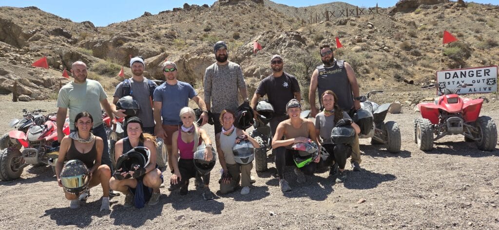 Group of 14 smiles with helmets by ATVs in Nevada desert near Las Vegas on an ATV tour; “DANGER STAY OUT” sign in view.