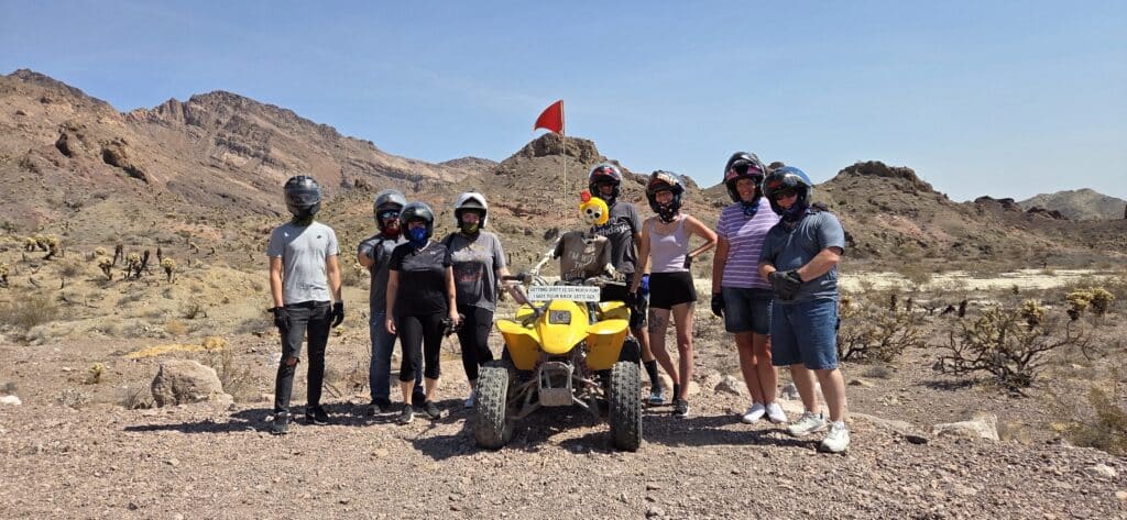 Nine people in helmets gather by a yellow ATV on a Nevada desert tour near Las Vegas with mountains and clear blue sky.