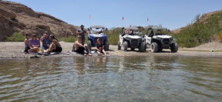 Friends relax by the Colorado River near Las Vegas, with RZR ATVs on a rocky shore, desert hills and shrubs in Nevada.