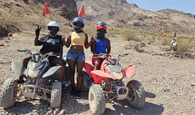 Three people on ATVs enjoy a Nevada desert tour near Las Vegas, posing with peace signs against mountain views.