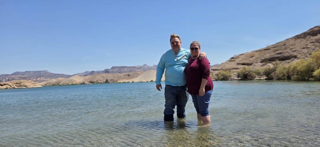 Smiling couple wades in the clear Colorado River near Las Vegas, Nevada, with rocky hills, blue sky, and sunglasses.