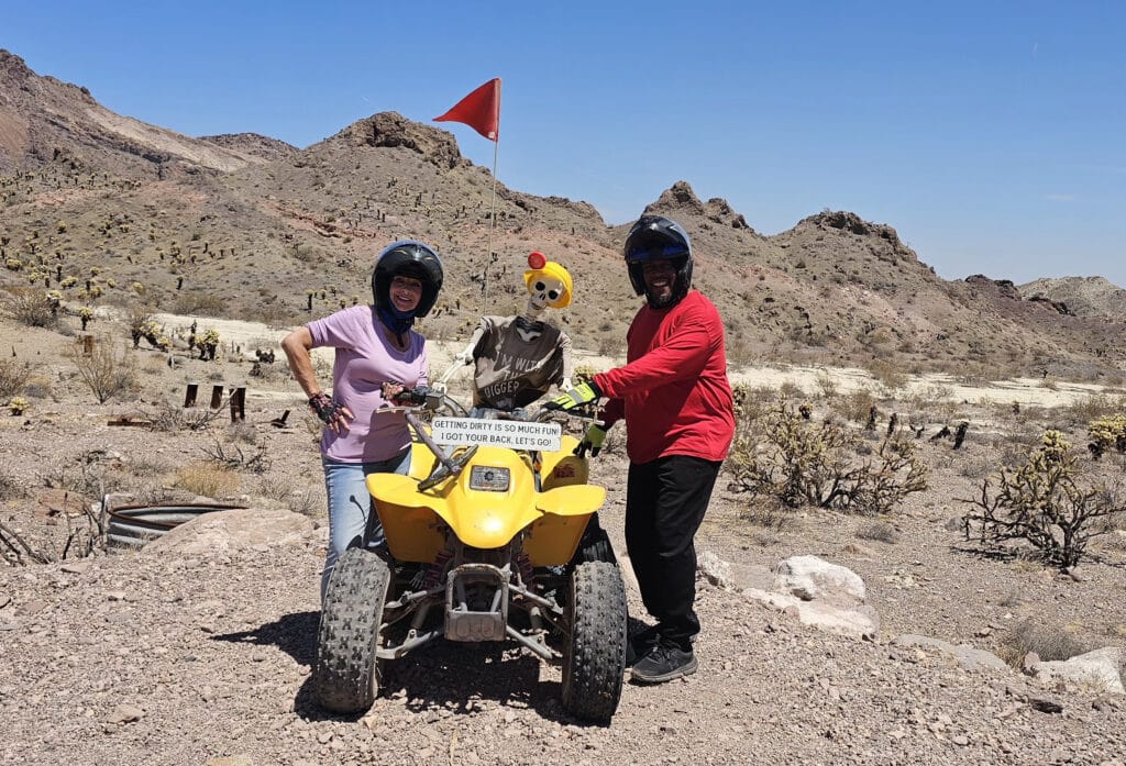 Two people with helmets pose by a yellow ATV in the Nevada desert; skeleton holds sign. Red flag, rocky Las Vegas hills behind.