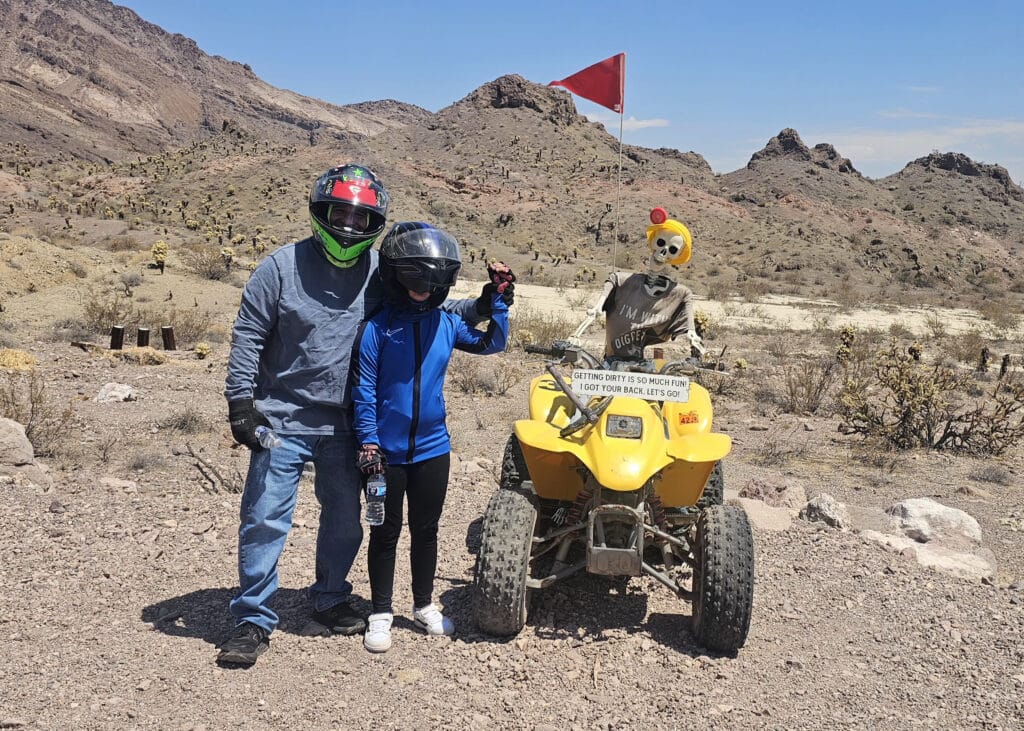 ATV tour near Las Vegas: two riders by a yellow ATV with a helmeted skeleton in Nevada’s desert mountains under blue sky.