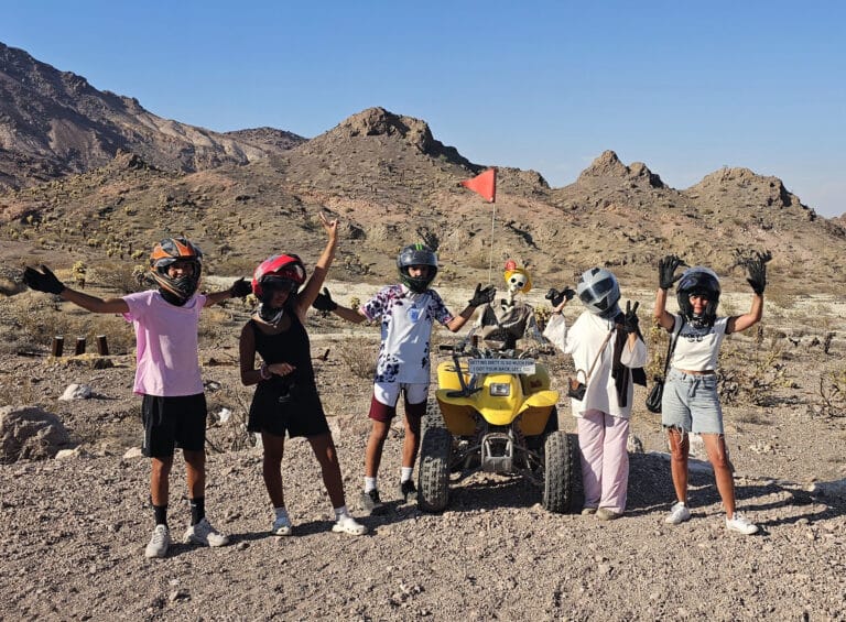 Group posing by yellow ATV on Nevada desert tour near Las Vegas, with helmets, gloves, and Rocky Mountains under clear blue sky.