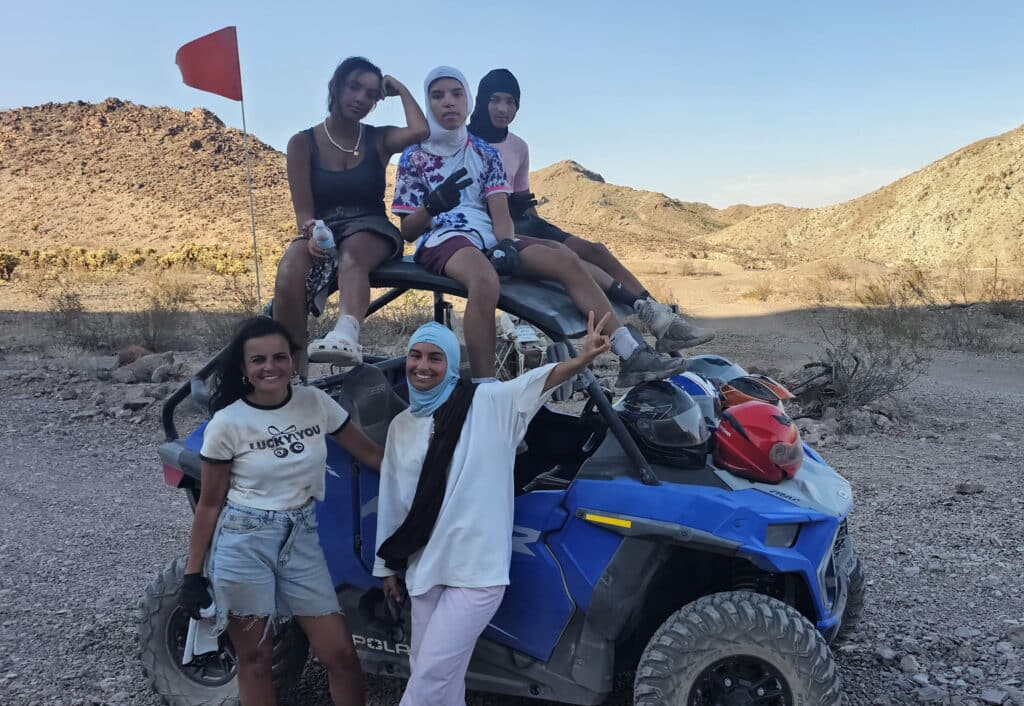 Five women enjoy an ATV tour near Las Vegas, posing on a blue RZR in a Nevada desert with mountains and a red safety flag.